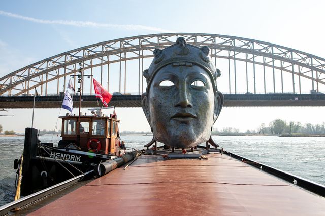 Het levensgrote masker staat op een aak, op de achtergrond de brug over de Waal.