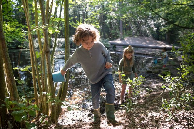 Een jongen en twee meisje spelen in met water, emmertjes en visnetjes bij een vennetje in één van de Drentse bossen.