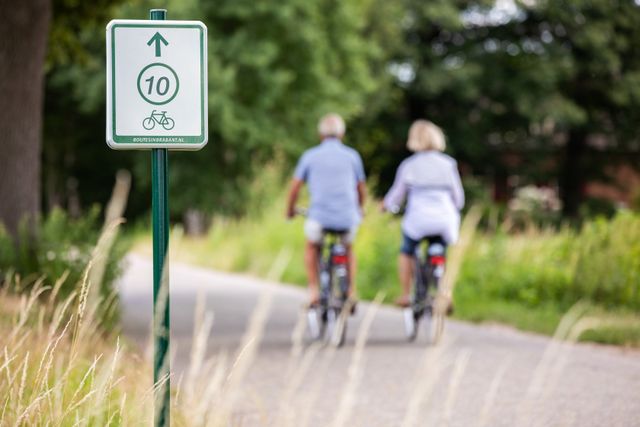 Een man en vrouw fietsen door de natuur. Hierbij is een bord met knooppunt in beeld.