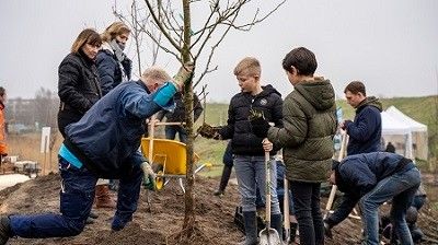Kinderen die een meneer helpen met het planten van een boom op een grondstuk met in de achtergrond moeders die aan het kijken zijn.