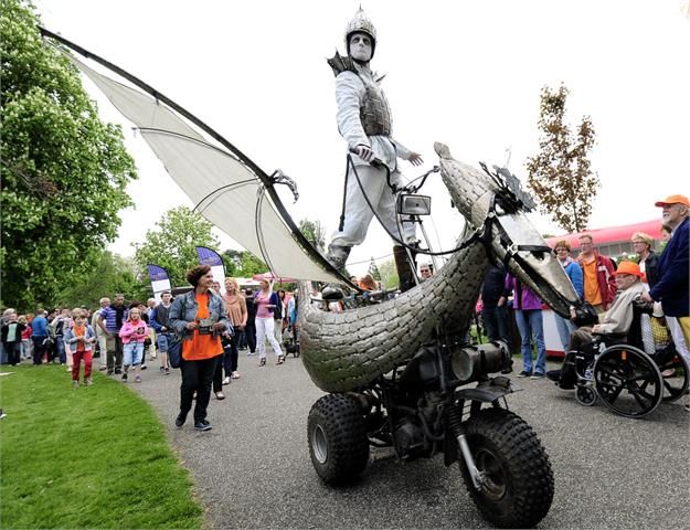 Een man die verkleed als draak is rijd met een elektrische fiets die ook opgemaakt is als draak en hier staan veel mensen en kinderen naar te kijken.