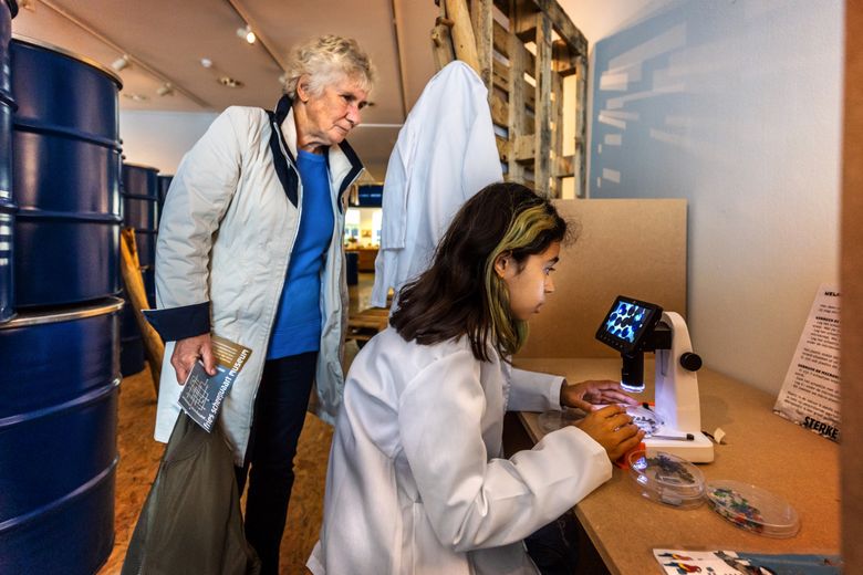 Een meisje in een witte laboratoriumjas aan kijkt door een microscoop. Een oudere vrouw kijkt toe.