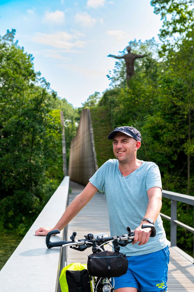 Een man met een fiets staat op een brug. Op de achtergrond zie je een hoge wal met daar doorheen een looppad en bovenop een kunstwerk van takken.