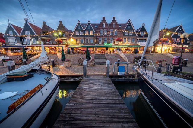 Een sfeerfoto van hotel Old Dutch genomen vanaf de haven met lichtjes en houten schepen in beeld